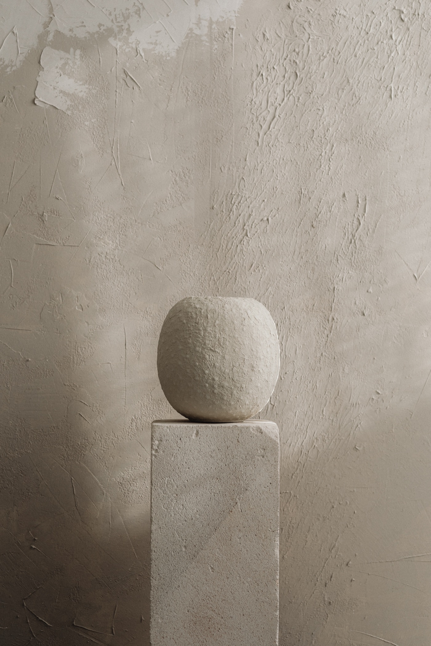 Round plaster vessel on a stone plinth against a hand-troweled plaster wall in soft window light