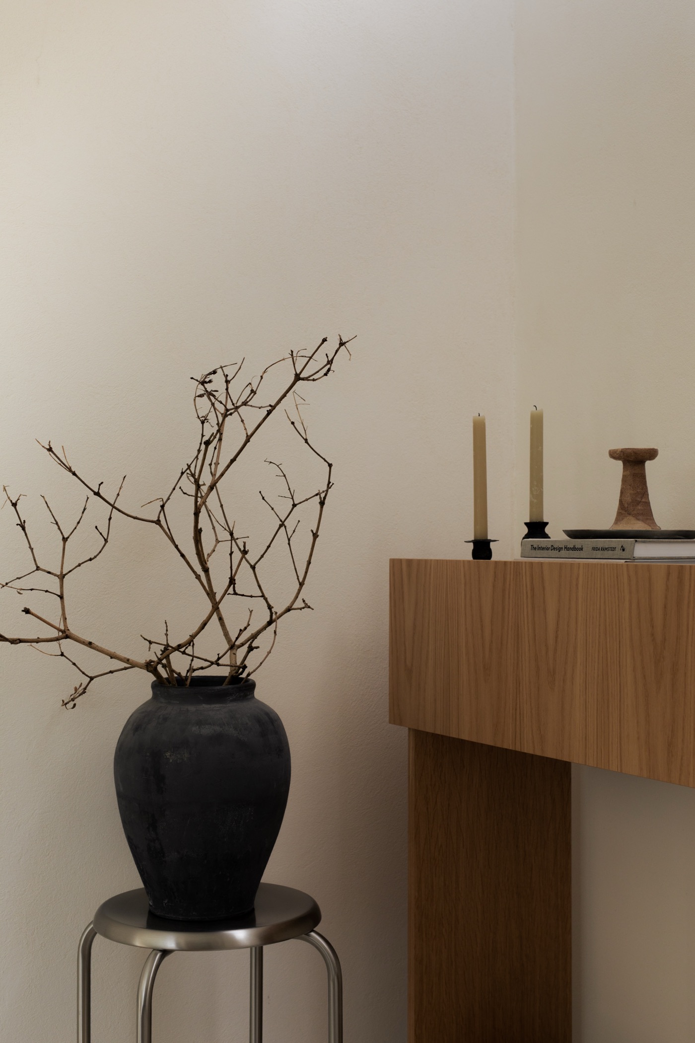 Black clay vase with bare branches on a steel stool beside an oak console with candles