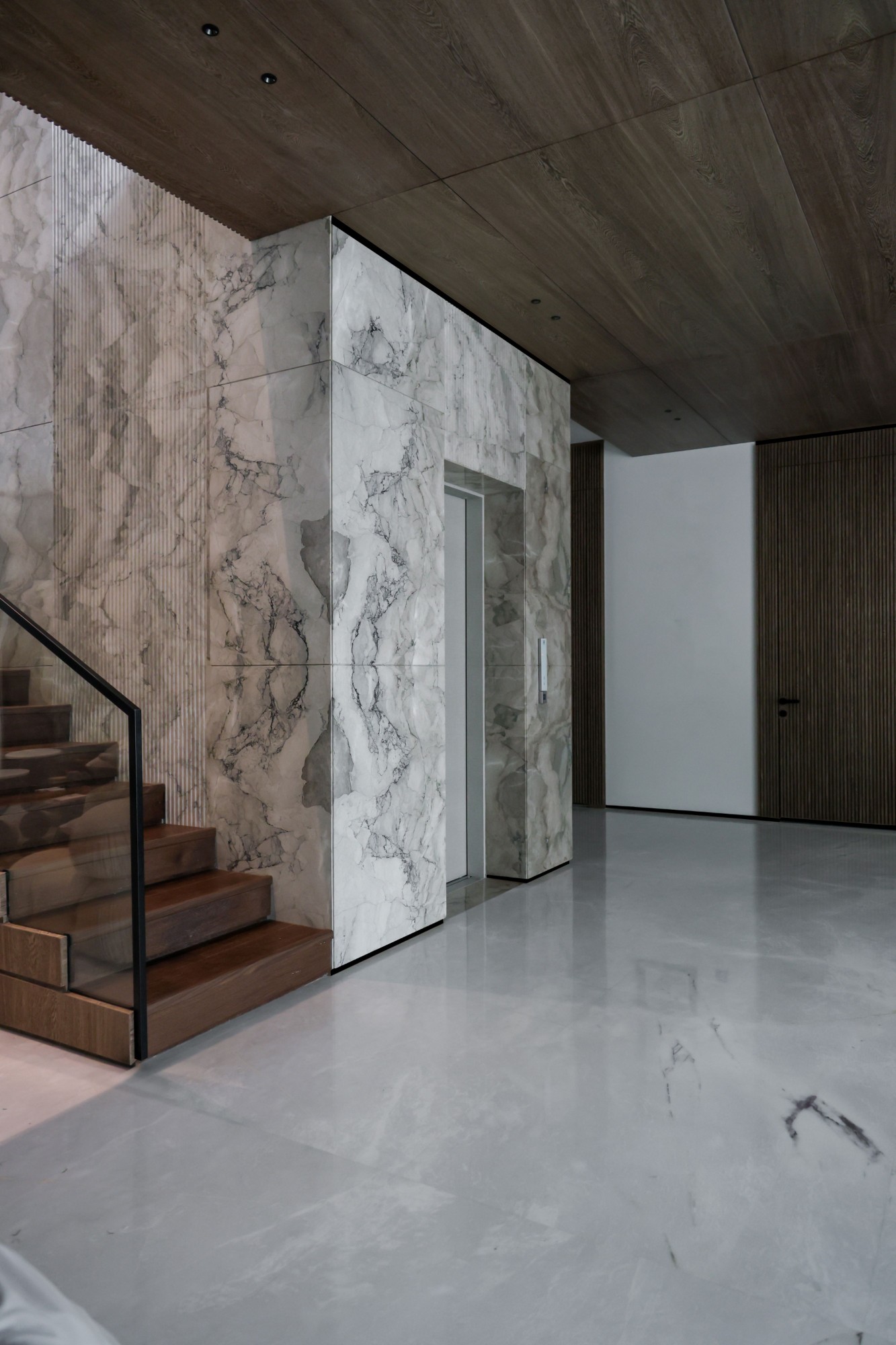 Book-matched marble wall and elevator beside an oak stair with glass balustrade