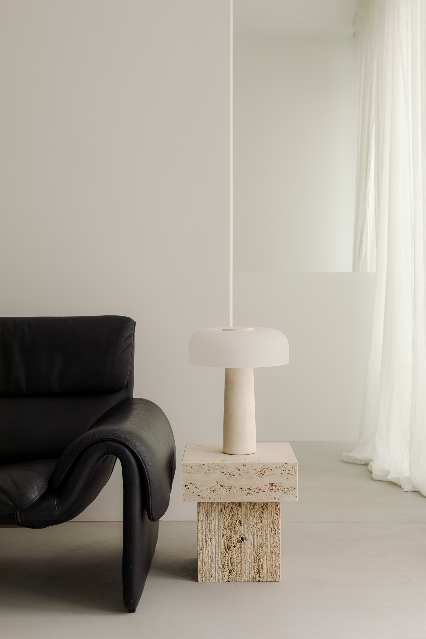 Black leather sofa beside a travertine side table and mushroom lamp, sheer linen curtains behind