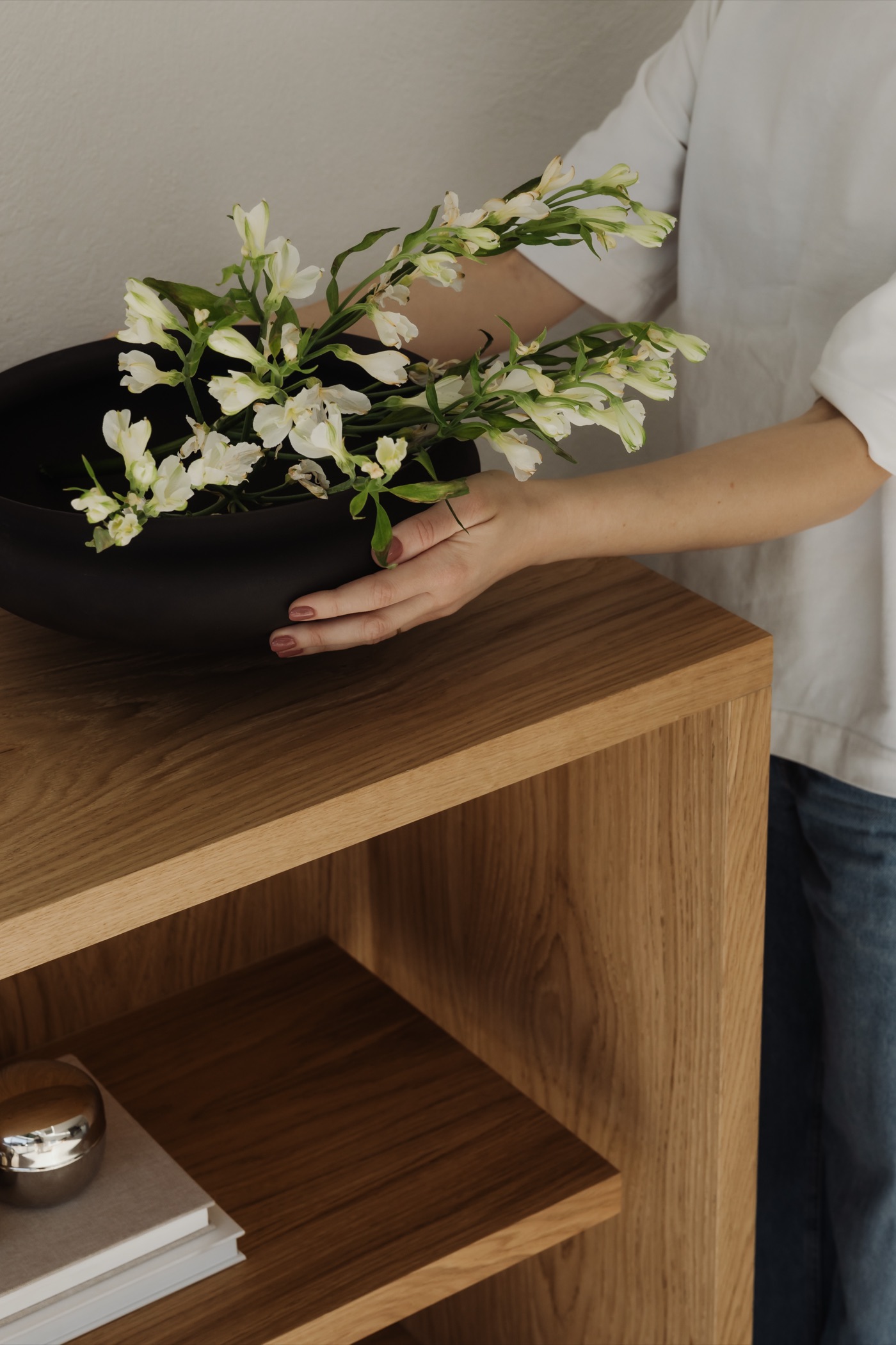 Hands arranging white flowers in a black ceramic bowl on an oak cabinet