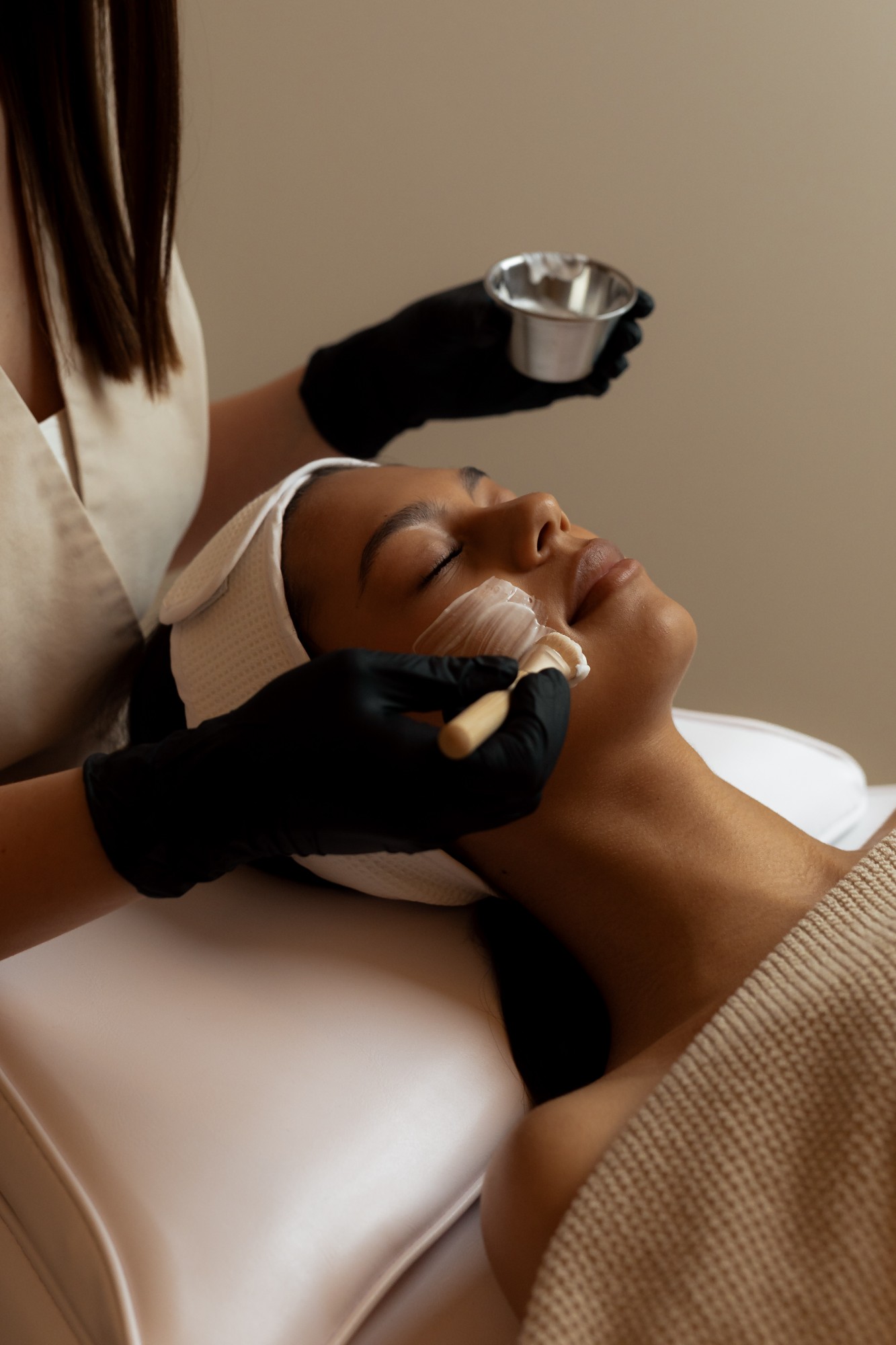 Esthetician in black gloves brushing a mask along a client’s jaw