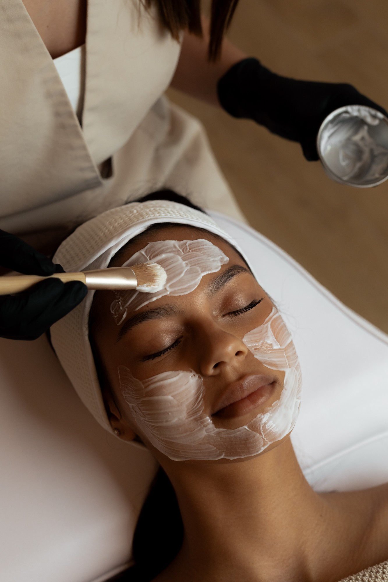 Esthetician brushing a cream mask onto a client lying with eyes closed